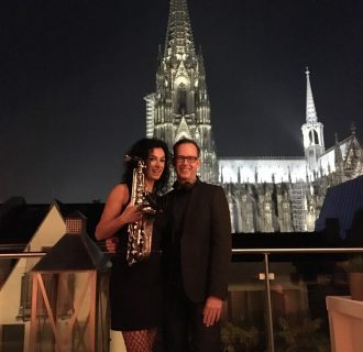 DJ René Pera und Angela Puxi. Als PuxiPera. DJ plus Saxophon. Sie stehen auf der Terrasse der Früh Lounge. Im Hintergrund sieht man den Kölner Dom.