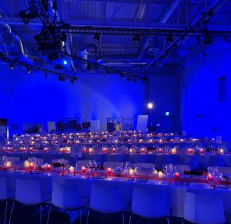 Top Event DJ René Pera bei einer Benefiz-Gala der AIDS Stiftung im Dock.One/Köln. Man blickt auf gedeckte, längliche Tafeln mit Kerzenlicht. Im Hintergrund sieht man einige Kunstwerke. Der Raum ist blau ausgeleuchtet.